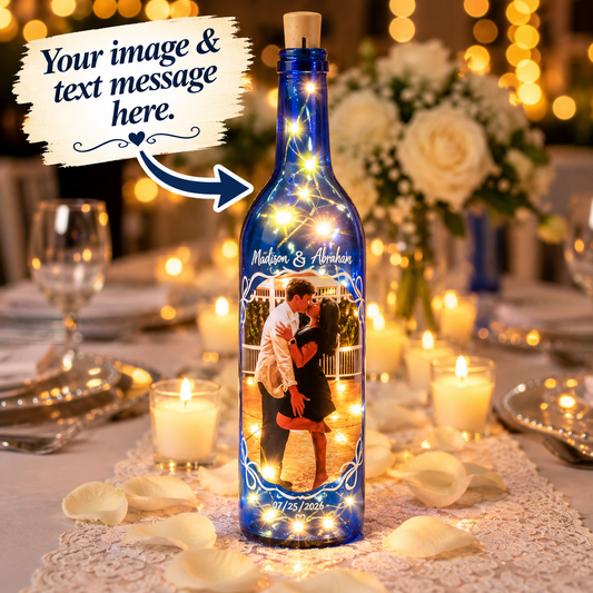 A personalized blue glass wine bottle with a cork stopper, filled with warm fairy lights and featuring a printed photo of a couple, set on a decorated dining table with candles, rose petals, and a blurred background. A graphic overlay at the top left says 'Your image & text message here.