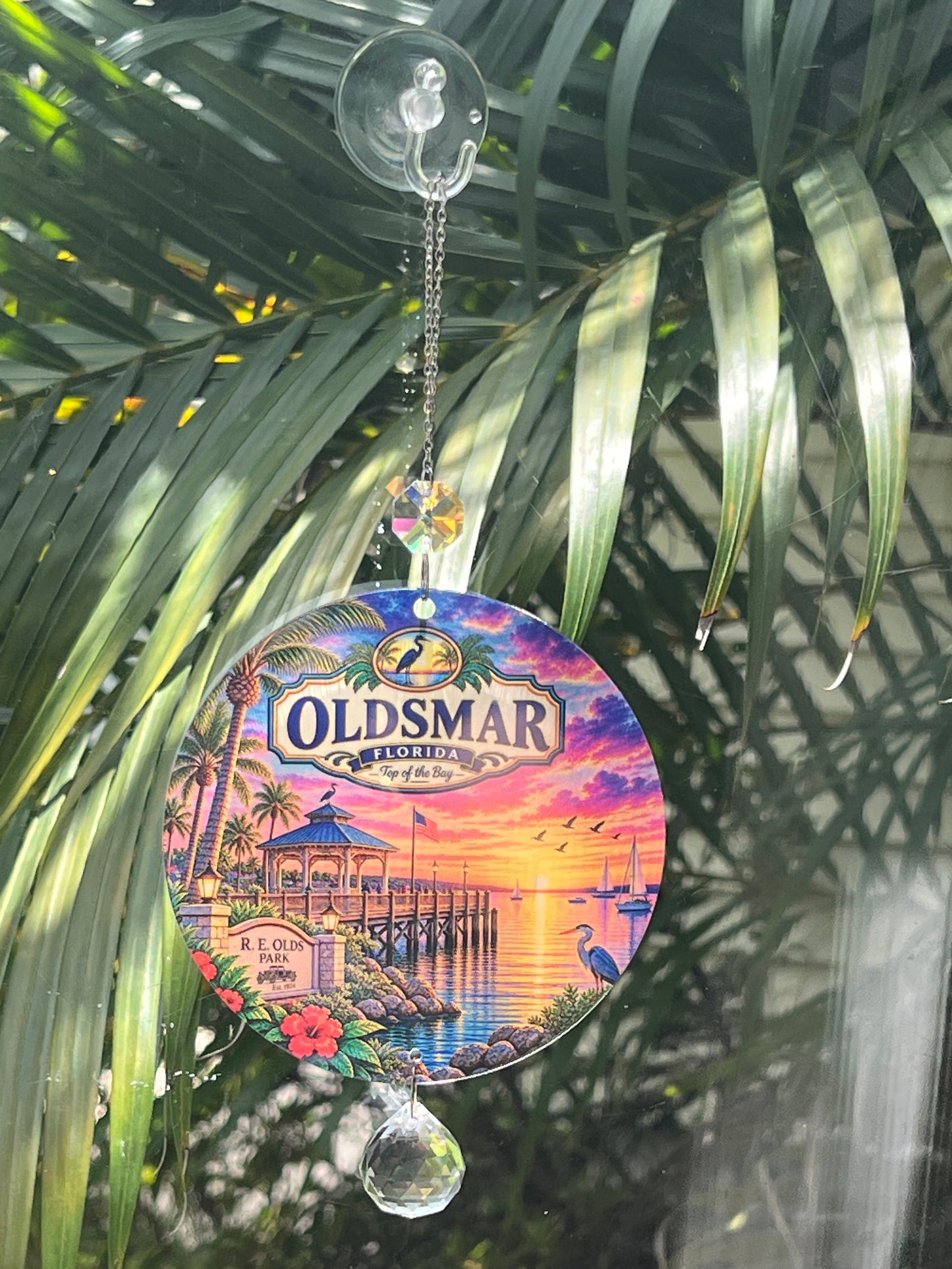 A circular Oldsmar Beach Boardwalk glass wind chime ornament hanging from a clear suction cup hook. The ornament features a colorful sunset beach boardwalk scene with palm trees, a pier, and a lighthouse, and is shown hanging in front of green palm leaves.