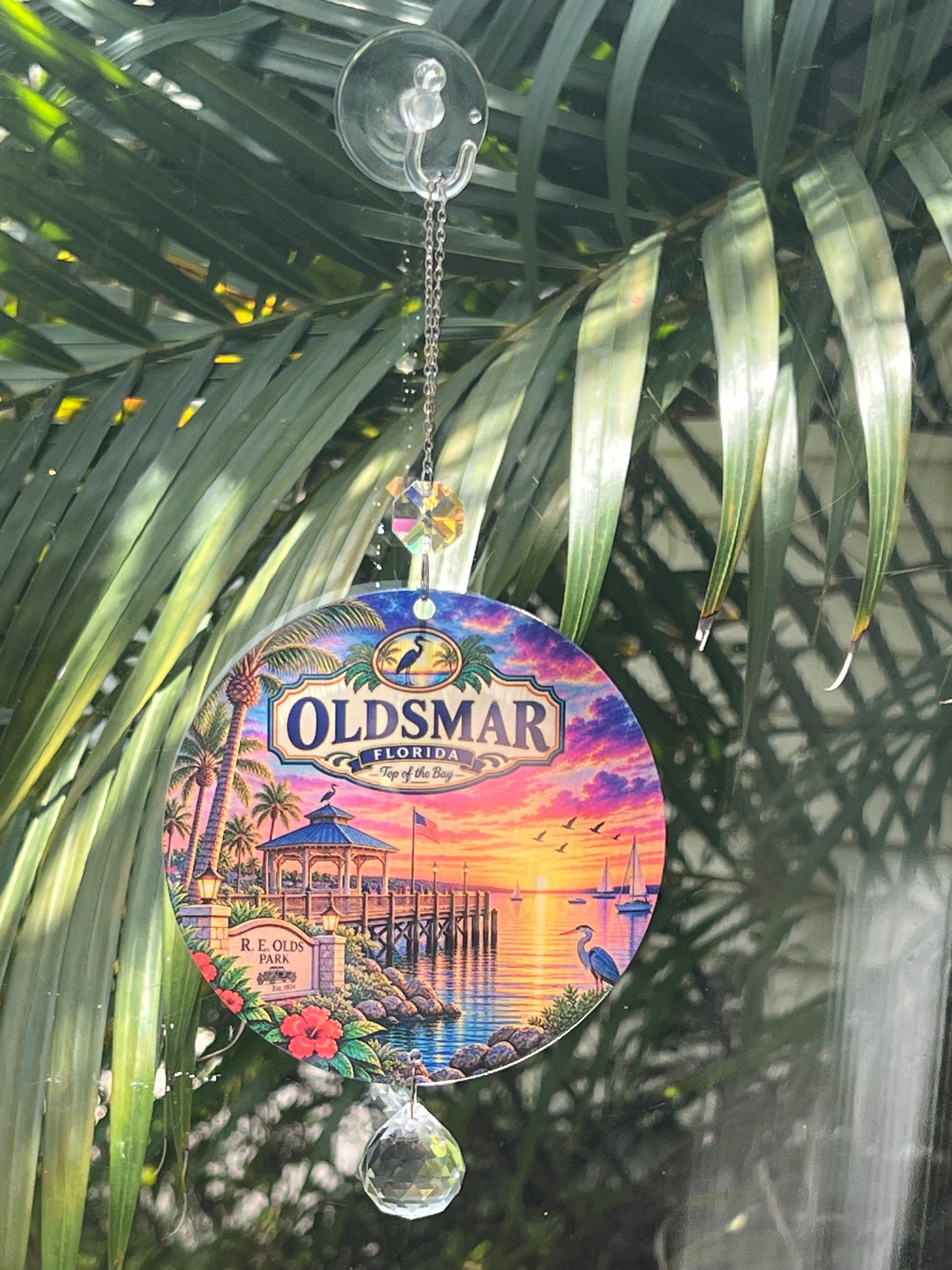A circular Oldsmar Beach Boardwalk glass wind chime ornament hanging from a clear suction cup hook. The ornament features a colorful sunset beach boardwalk scene with palm trees, a pier, and a lighthouse, and is shown hanging in front of green palm leaves.