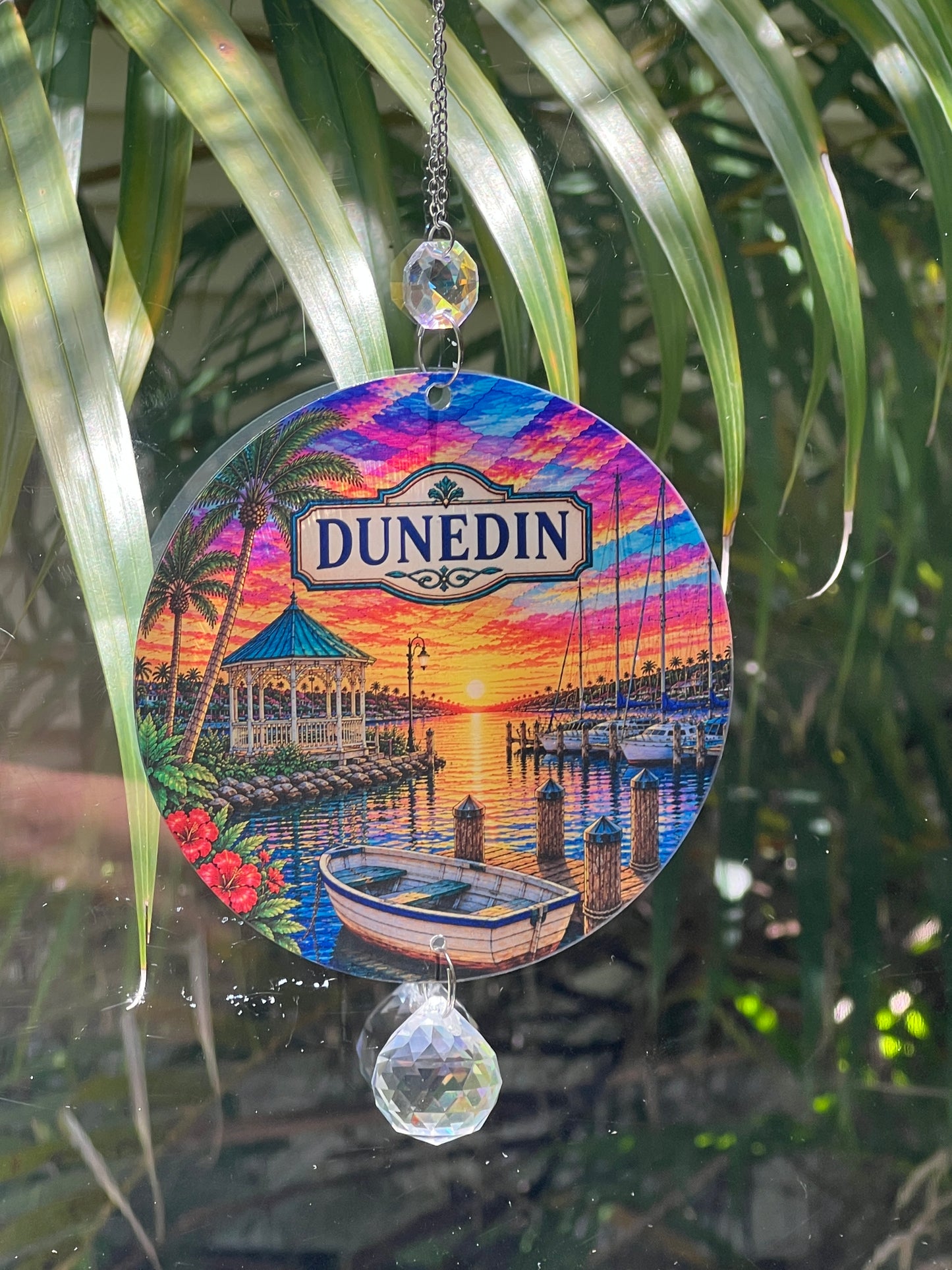 A circular Dunedin-themed sun gazing ornament hanging in front of palm leaves. The ornament features a colorful sunset marina scene with a boat, dock, palm trees, and a gazebo, with the 'DUNEDIN' logo at the top.