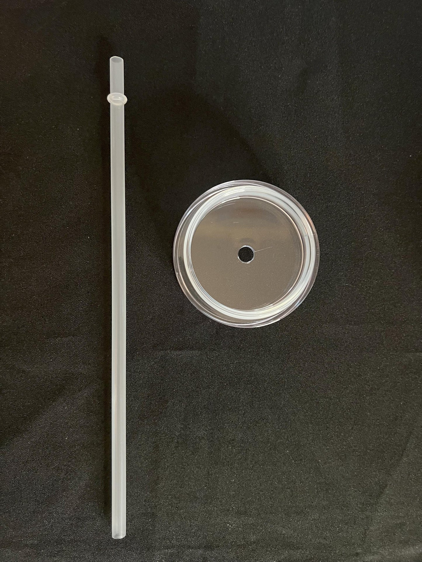 A clear plastic straw and a clear plastic lid with a metal rim, displayed on a black fabric background.