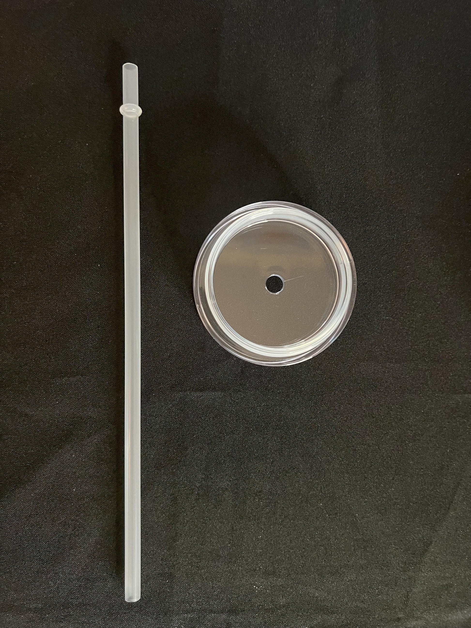 A clear plastic straw and a clear plastic lid with a metal rim, displayed on a black fabric background.