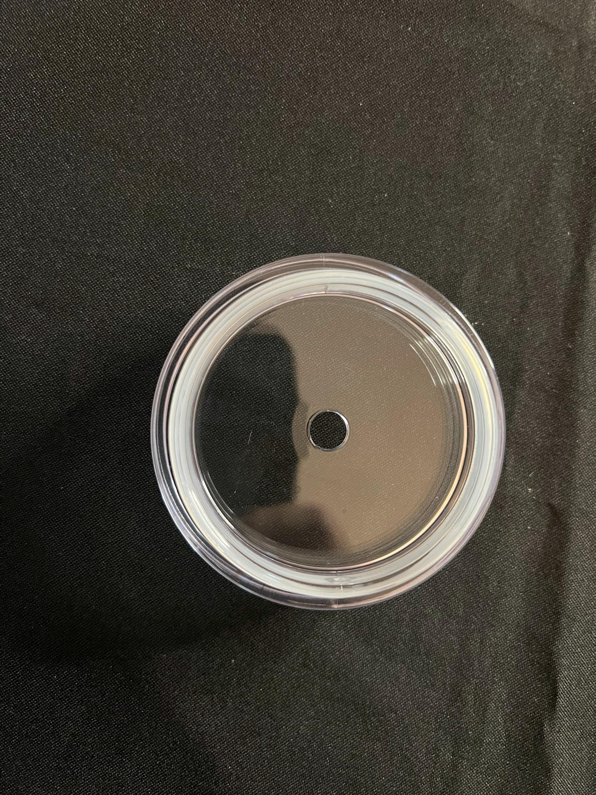 A clear plastic lid with a single central hole, shown from a top-down view on a black textured fabric background.