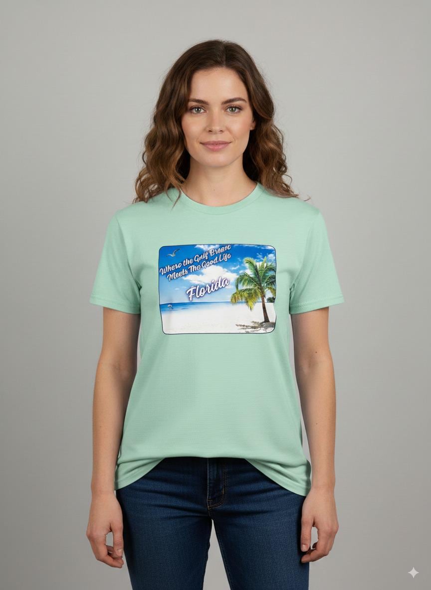 Person wearing a mint green 'Florida' t-shirt on a beach with a sailboat and palm trees in the background. T-shirt has a  picture on the front of white sand beach, aqua gulf waters, palm tree and the text "where the gulf breeze meets the good life" Florida. 