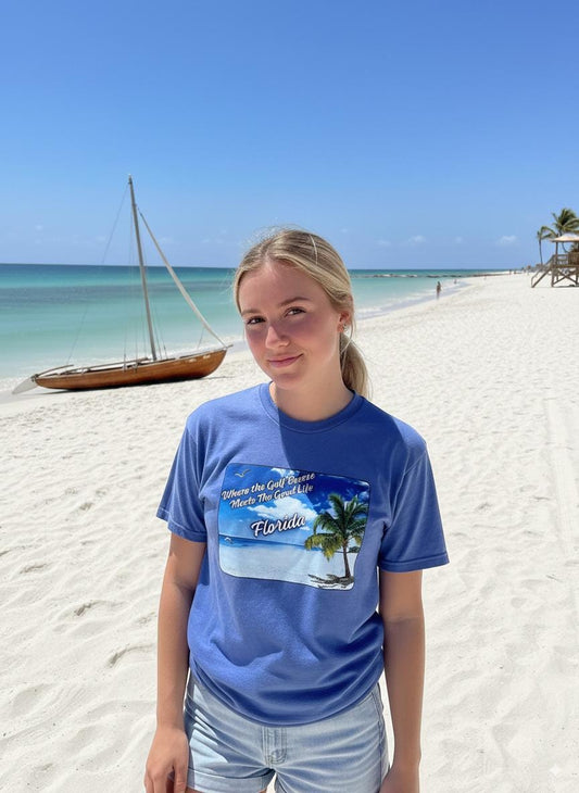 Person wearing a purple 'Florida' t-shirt on a beach with a sailboat and palm trees in the background. T-shirt has a  picture on the front of white sand beach, aqua gulf waters, palm tree and the text "where the gulf breeze meets the good life" Florida. 